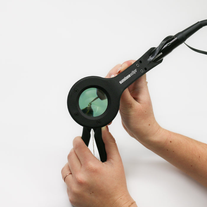 Pictured is two hands holding permanent jewelry (PJ) chain with grounded precision pliers and also holding a PJ welder stylus. This accessory is attached to the stylus and a weld is being demonstrated on the chain underneath the magnification lens.