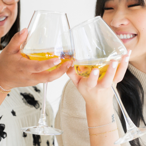 two women wearing permanent jewelry clink wine classes together to celebrate their success with permanent jewelry