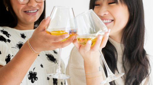 two women wearing permanent jewelry clink wine classes together to celebrate their success with permanent jewelry