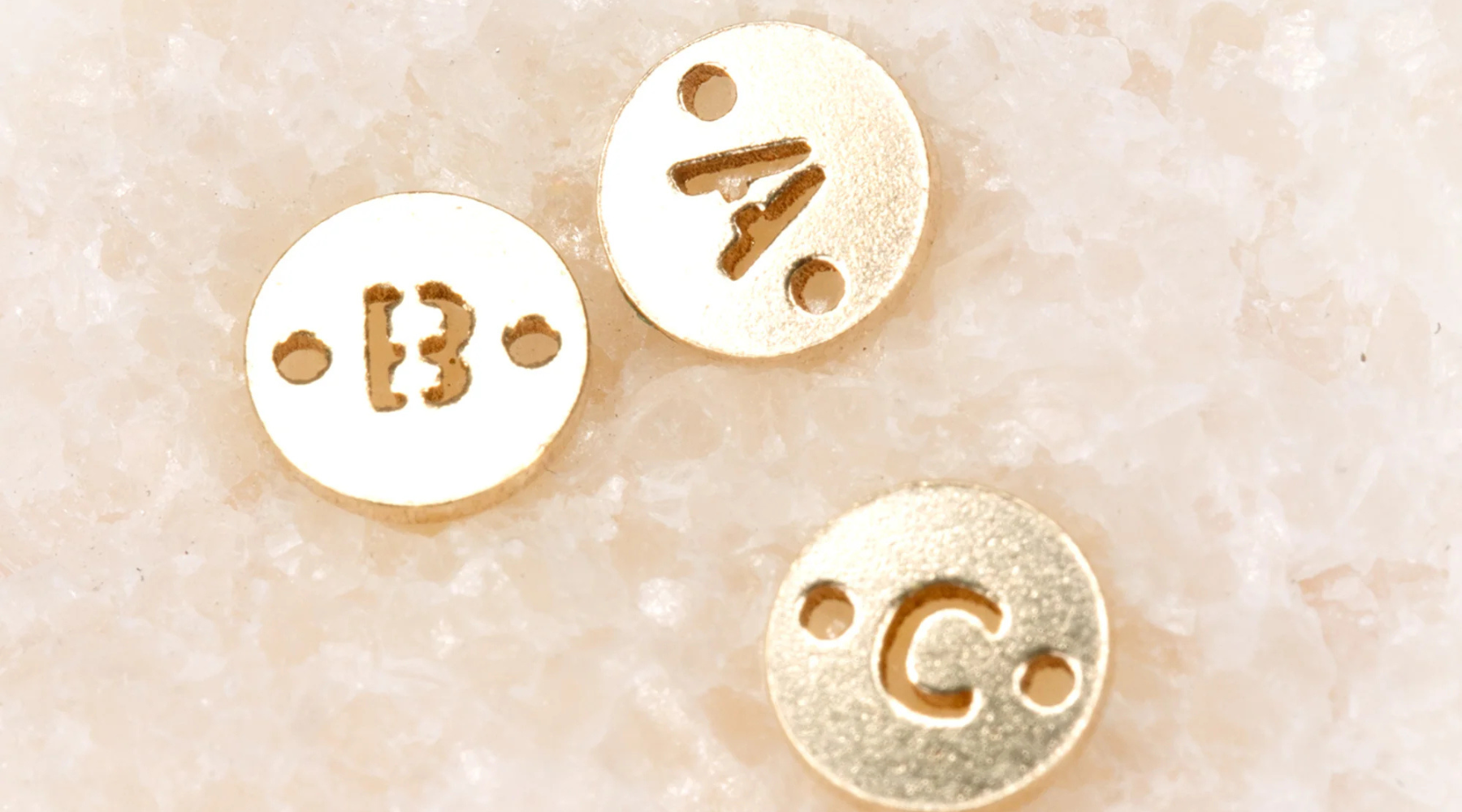 An image of three disc initial connectors used for permanent jewelry. They each have a letter of the alphabet in the middle (A, B, and C) and have two holes in the side to connect to a bracelet or necklace.