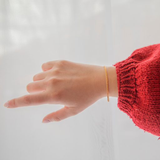 Hand wearing a gold interlinked permanent jewelry chain bracelet with a red sleeve on a light background