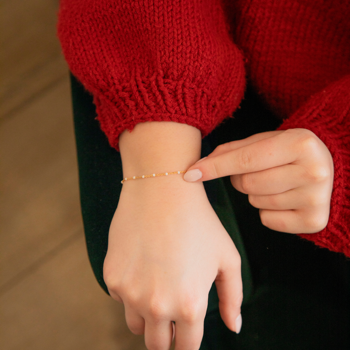 Person wearing a red sweater pointing to a gold enamel permanent jewelry bracelet chain on their wrist.