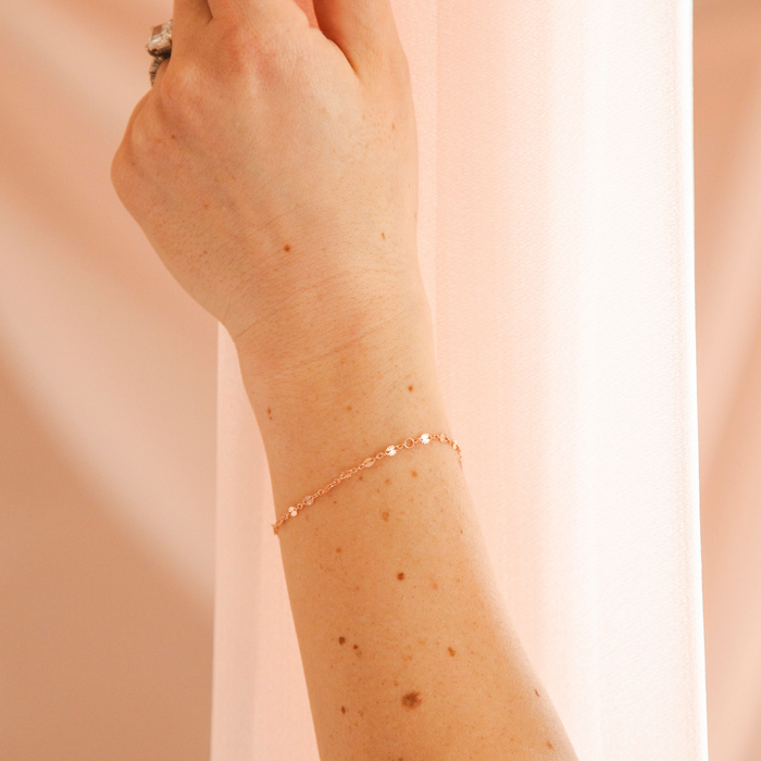 Photo is of a models hand in front of some soft pink curtains. The model is wearing a rose gold chain.