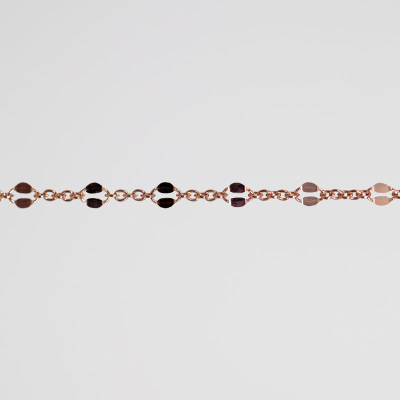 A close-up of a delicate rose gold chain bracelet featuring alternating hammered oval links in polished rose gold, set against a solid light gray background.