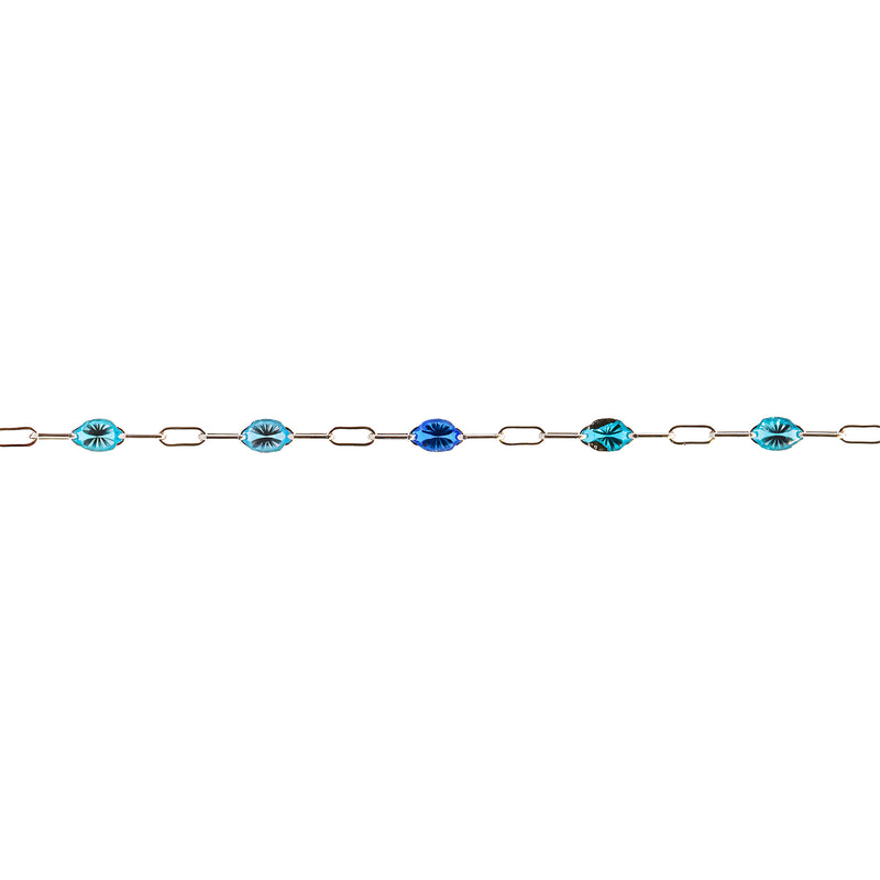 A delicate permanent jewelry chain featuring oval and elongated links, interspersed with enamel beads in shades of light blue, royal blue, and turquoise with dark inclusions, shown against a plain white background.
