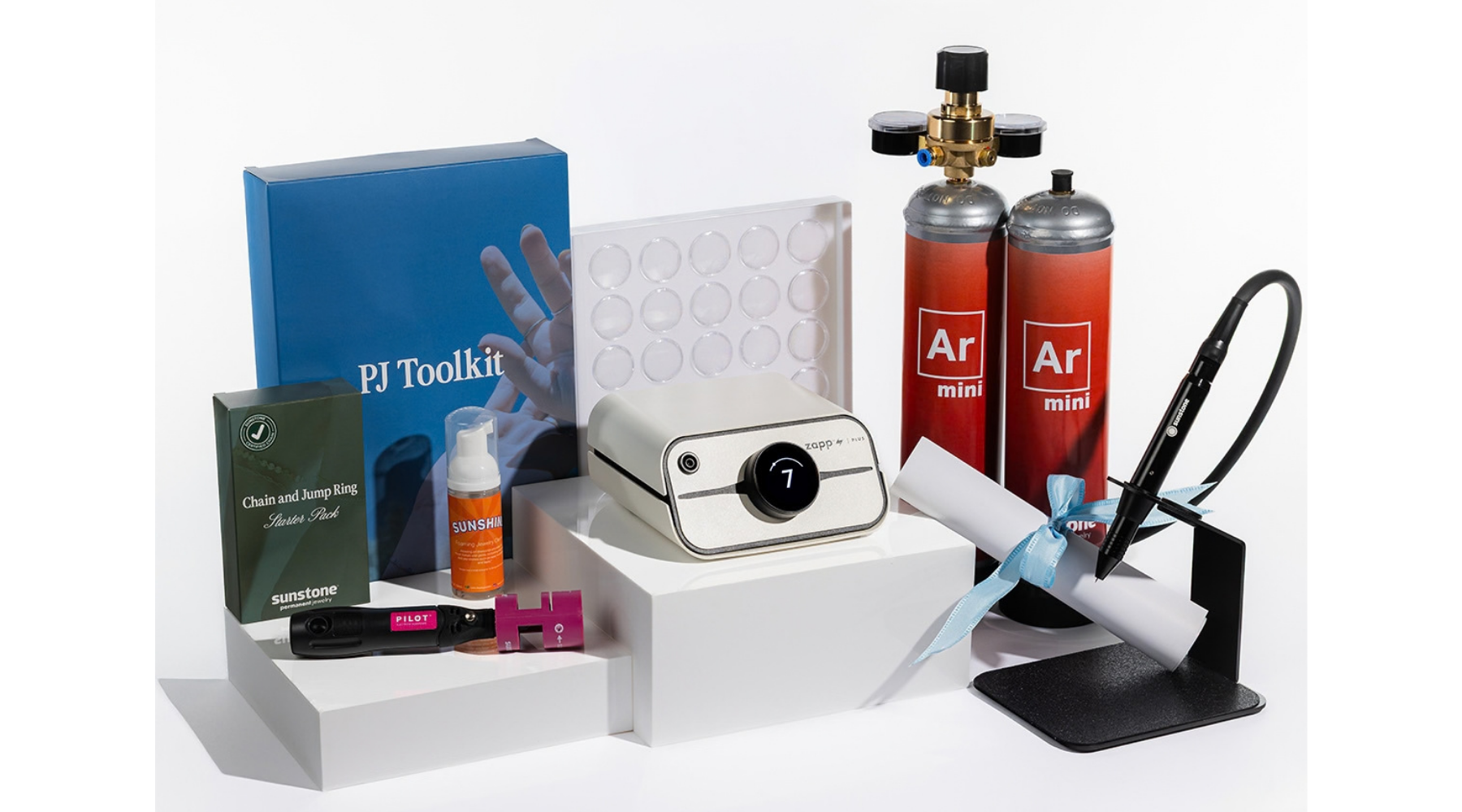 A PJ Toolkit kit arranged on white display blocks against a clean white background. The kit includes a white Zapp Plus 2 welder, two orange and red argon mini cylinders labeled 'Ar mini', a blue PJ Toolkit box, a small orange cleaner for PJ, a black and pink electrode sharpener, a white sheet on a stand with a blue ribbon, and other small accessories.