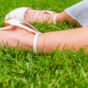A delicate gold chain anklet worn on a person's ankle, shown outdoors on green grass next to white flat sandals.