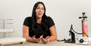 A woman with long black hair sits at a table wearing a black t-shirt, gesturing with her hands. On the table in front of her are various permanent jewelry tools, including a permanent jewelry welder, a small display of pj chains, and a pink argon gas cylinder.