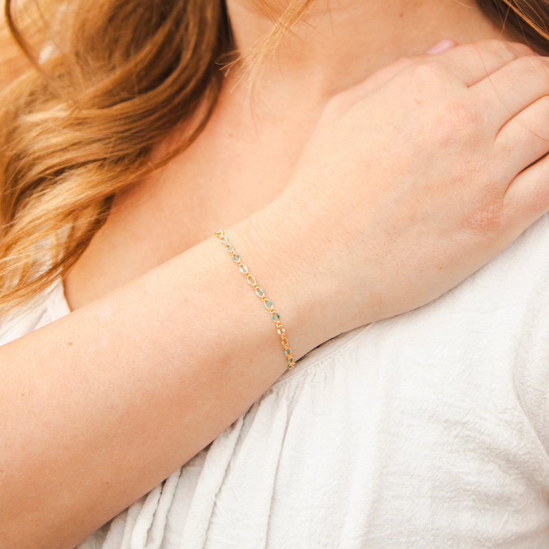 Model's hand is on chest showcasing a gold chain with blue, green, and white enamel beads. 