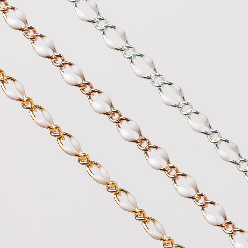 Zoomed in photo of three chains with white enamel on a gray background. One chain is gold, one is rose gold, and one is silver.