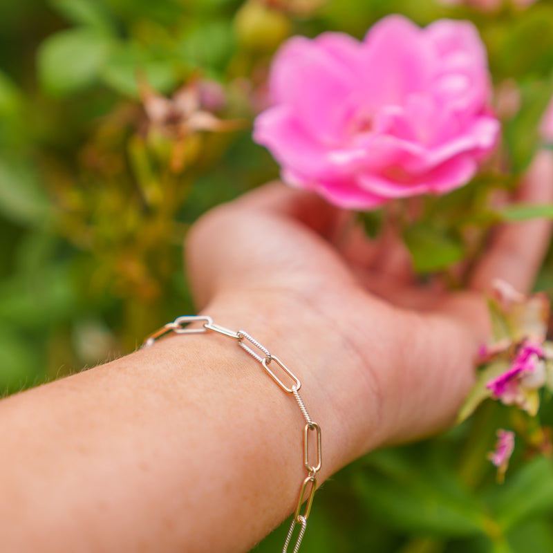 A close-up of a person's hand wearing a delicate gold and silver paperclip chain bracelet, set against a soft-focus background of green foliage and a pink rose.