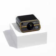 Black and gold Zapp Plus permanent jewelry welder on a square white pedestal casting a shadow on a white background