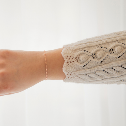 Close-up of a womans arm wearing a beige knit sweater with a subtle pattern and a paperclip permanent jewelry chain on a white background.