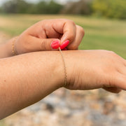 A close-up lifestyle shot of a delicate gold chain bracelet being adjusted on a person's wrist, with a blurred outdoor park background.