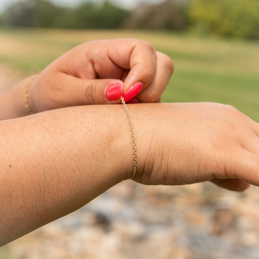 A close-up lifestyle shot of a delicate gold chain bracelet being adjusted on a person's wrist, with a blurred outdoor park background.