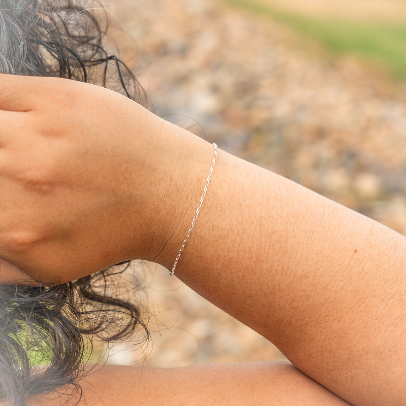 A delicate silver chain bracelet worn on a person's wrist, shown in a close-up outdoor shot with a blurred background of pebbles and greenery.