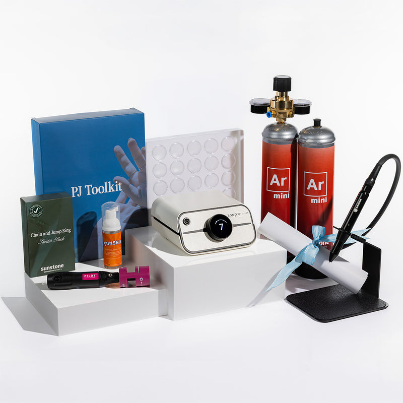 A PJ Toolkit kit arranged on white display blocks against a clean white background. The kit includes a white Zapp Plus 2 welder, two orange and red argon mini cylinders labeled 'Ar mini', a blue PJ Toolkit box, a small orange cleaner for PJ, a black and pink electrode sharpener, a white sheet on a stand with a blue ribbon, and other small accessories.