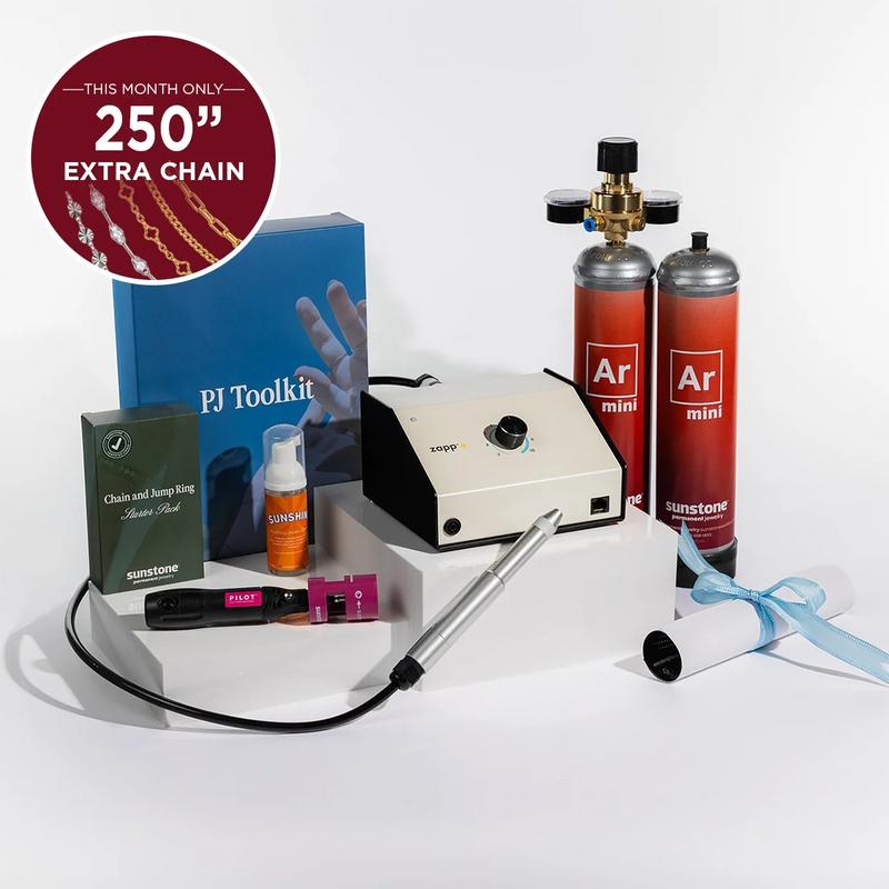 Permanent Jewelry tool kit arranged on white display blocks against a clean white studio background. The set includes a cream and black zapp welding machine, two red argon gas cylinders labeled Ar mini, a blue PI Toolkit box, a green chain and hoop ring box, an orange Sunstone jewelry cleaner spray bottle, a black and pink electrode sharpener, a silver welding stylus tool, a rolled white tube with a black cap representing training, and a blue ribbon.