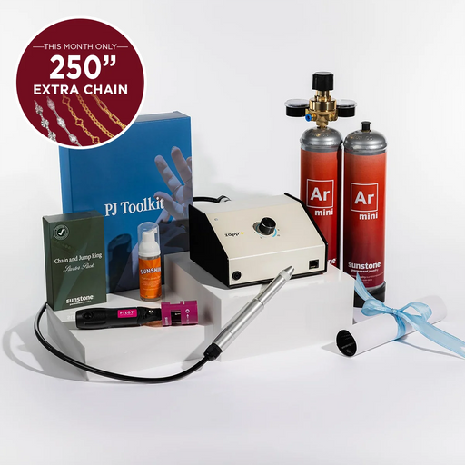 Permanent Jewelry tool kit arranged on white display blocks against a clean white studio background. The set includes a cream and black zapp welding machine, two red argon gas cylinders labeled Ar mini, a blue PI Toolkit box, a green chain and hoop ring box, an orange Sunstone jewelry cleaner spray bottle, a black and pink electrode sharpener, a silver welding stylus tool, a rolled white tube with a black cap representing training, and a blue ribbon.