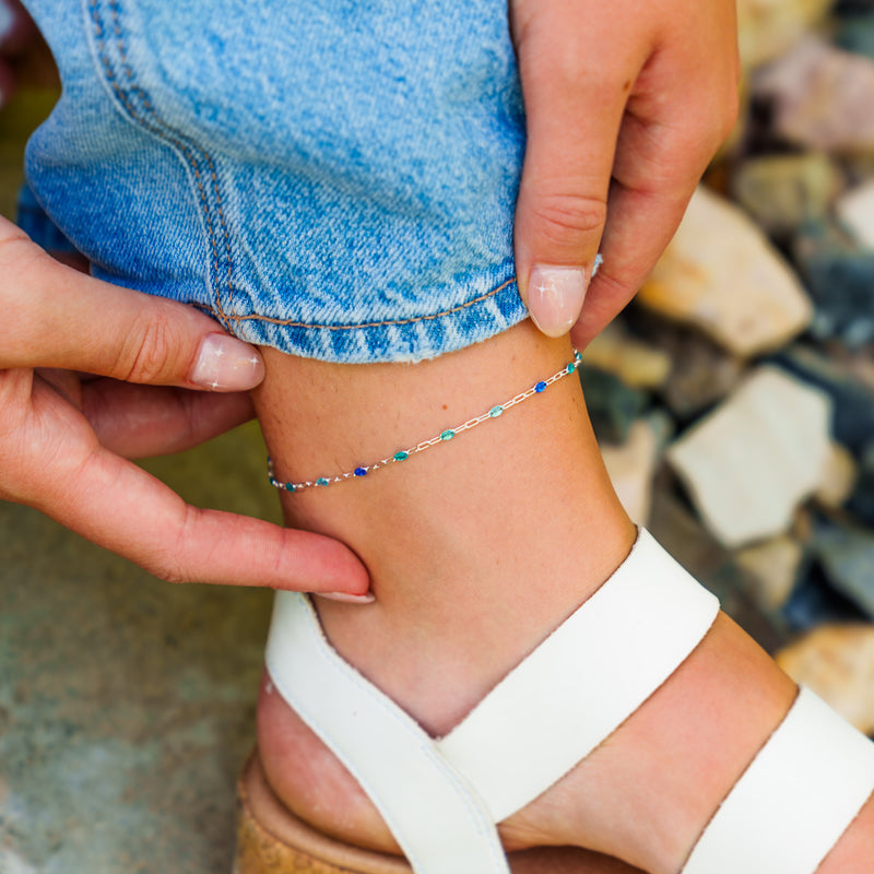 A detailed view of a silver permanent jewelry chain with tiny blue enamel beads of different shades worn as a delicate anklet.