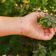 A lifestyle image of a person reaching for a plant, showcasing a silver permanent chain with different shades of purple enamel beads styled as a dainty bracelet.