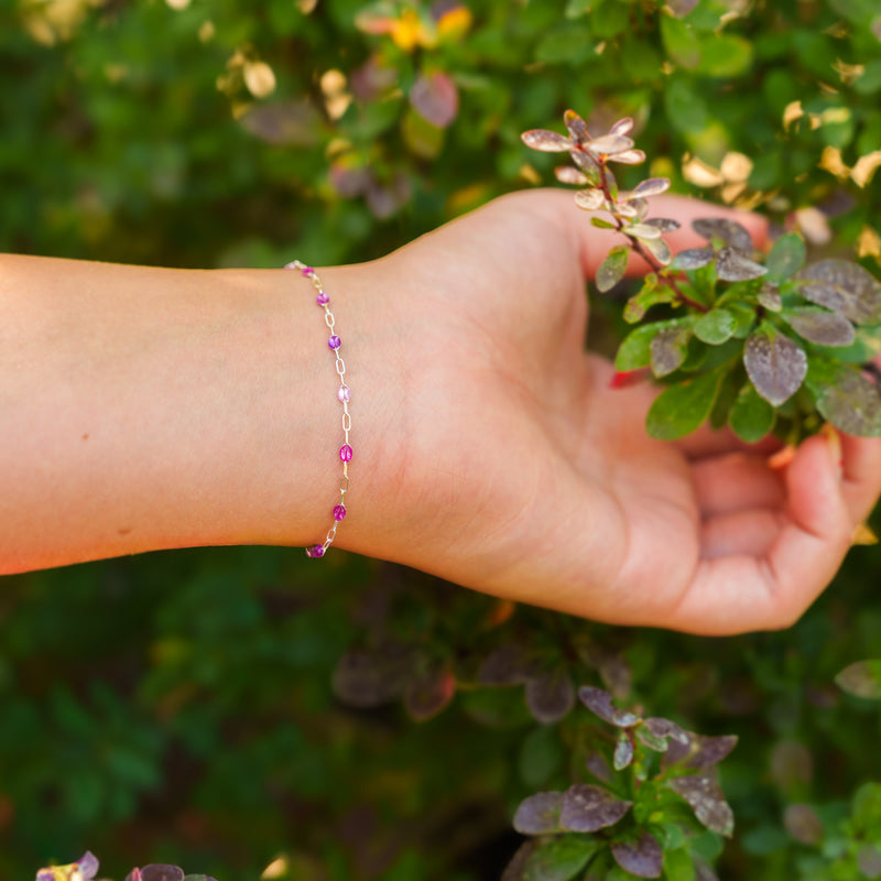 A lifestyle image of a person reaching for a plant, showcasing a silver permanent chain with different shades of purple enamel beads styled as a dainty bracelet.