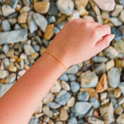 A lifestyle overhead photo of a fine gold curb link permanent jewelry chain worn as a bracelet against a background of river rocks.