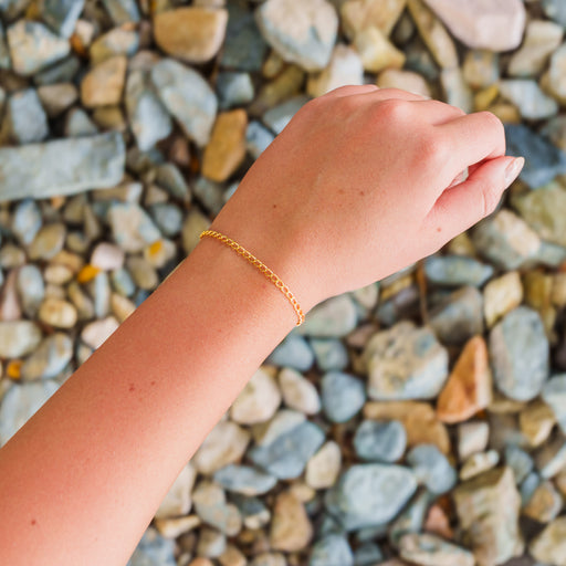A lifestyle overhead photo of a fine gold curb link permanent jewelry chain worn as a bracelet against a background of river rocks.