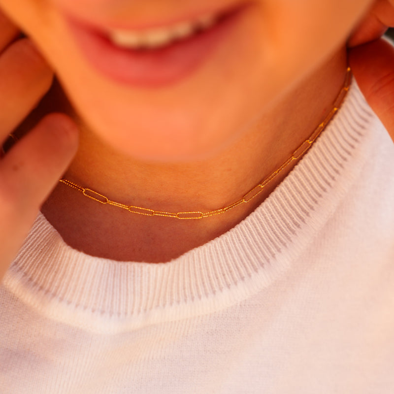 A lifestyle shot of a person smiling, showcasing a fine gold paperclip chain worn as a delicate choker-style permanent jewelry necklace.