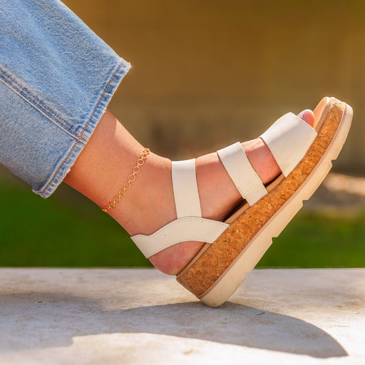 A lifestyle photo of a gold interlocking round link chain styled as a permanent jewelry anklet, worn with white wedge sandals.