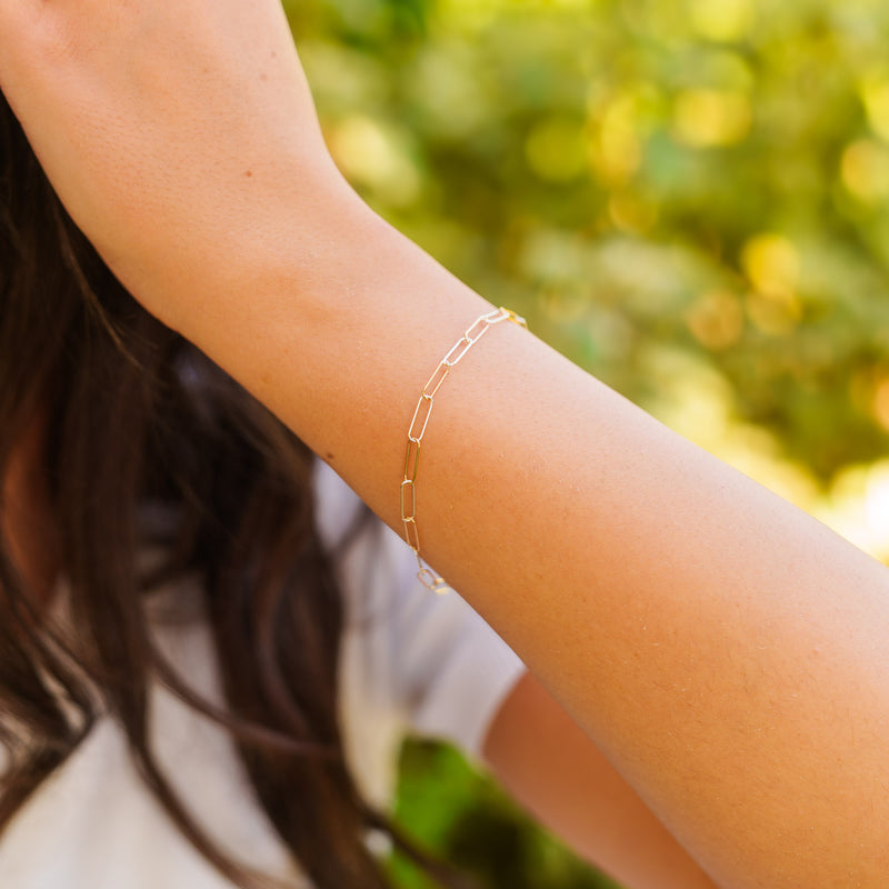 A lifestyle photograph showing a slender gold and silver mixed metal elongated paperclip permanent chain worn as a bracelet on a wrist with a soft natural green backdrop.
