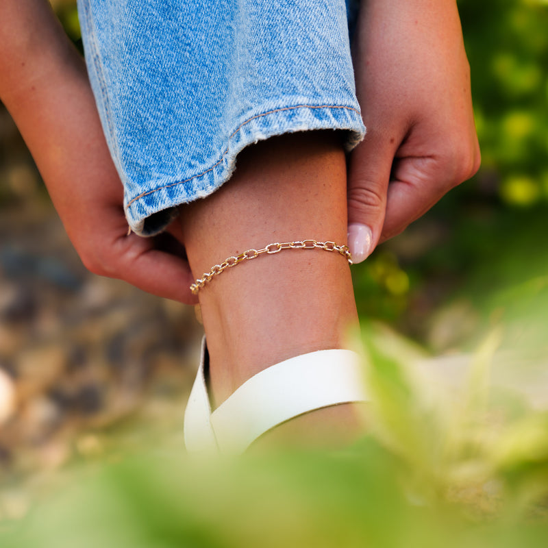 A detailed lifestyle image of a person's ankle showing the Nelly mixed-metal permanent jewelry chain worn as an anklet, paired with a white sandal strap.
